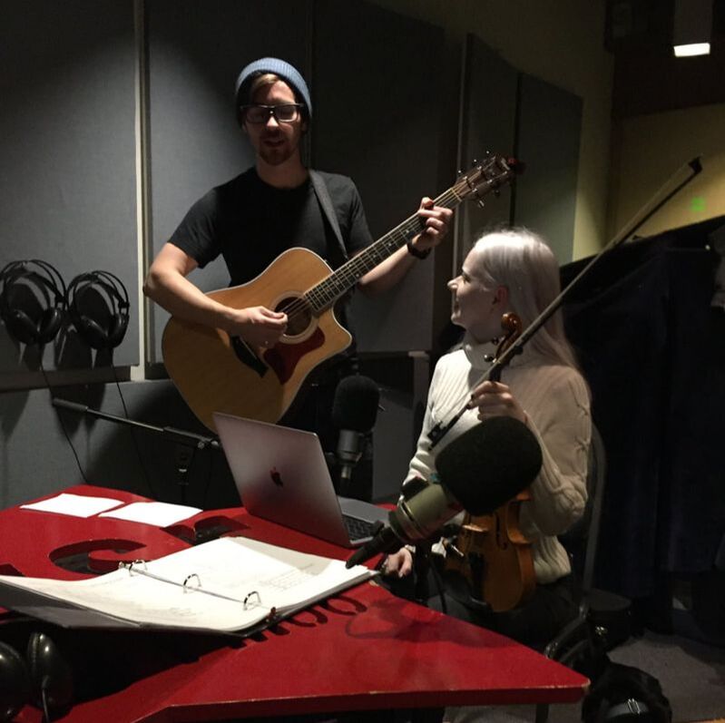 Tamara holding a violin in a radio station next to a man with a guitar