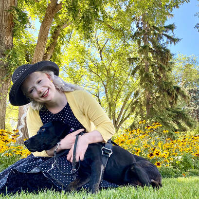 Tamara smiles and hugs her guide dog tight to her chest as they sit in the grass together. She is wearing a vintage 1950s polkadot swing dress with matching hat and a yellow cardigan.